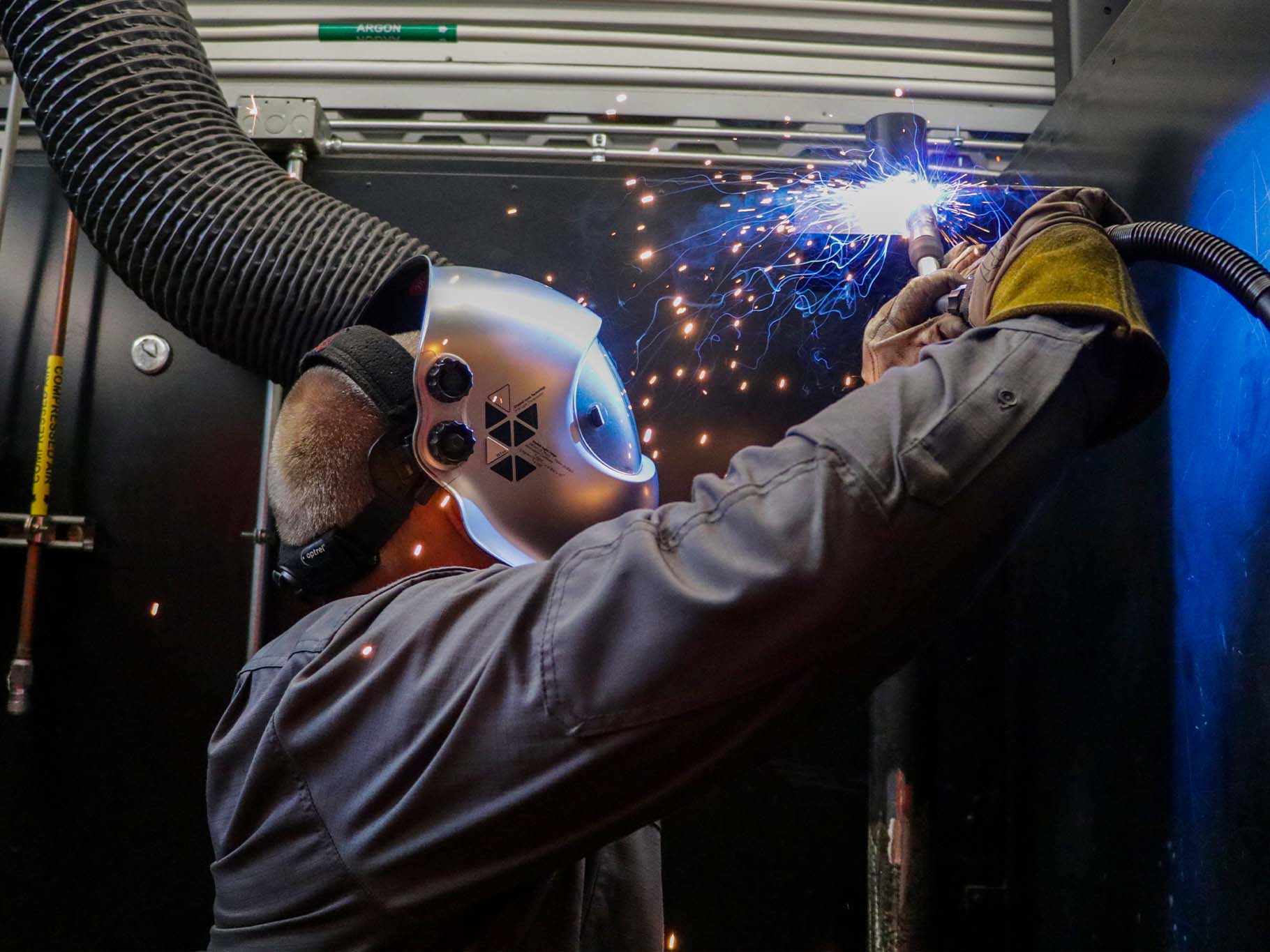 A worker wearing a protective welding helmet and gloves welds a metal surface indoors, with bright blue sparks flying from the welding torch and a ventilation hose visible overhead.