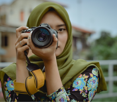 digial photography woman with a green scarf on her head and neck with a black blouse and blue floral patter holding a digital camera with a wide lens